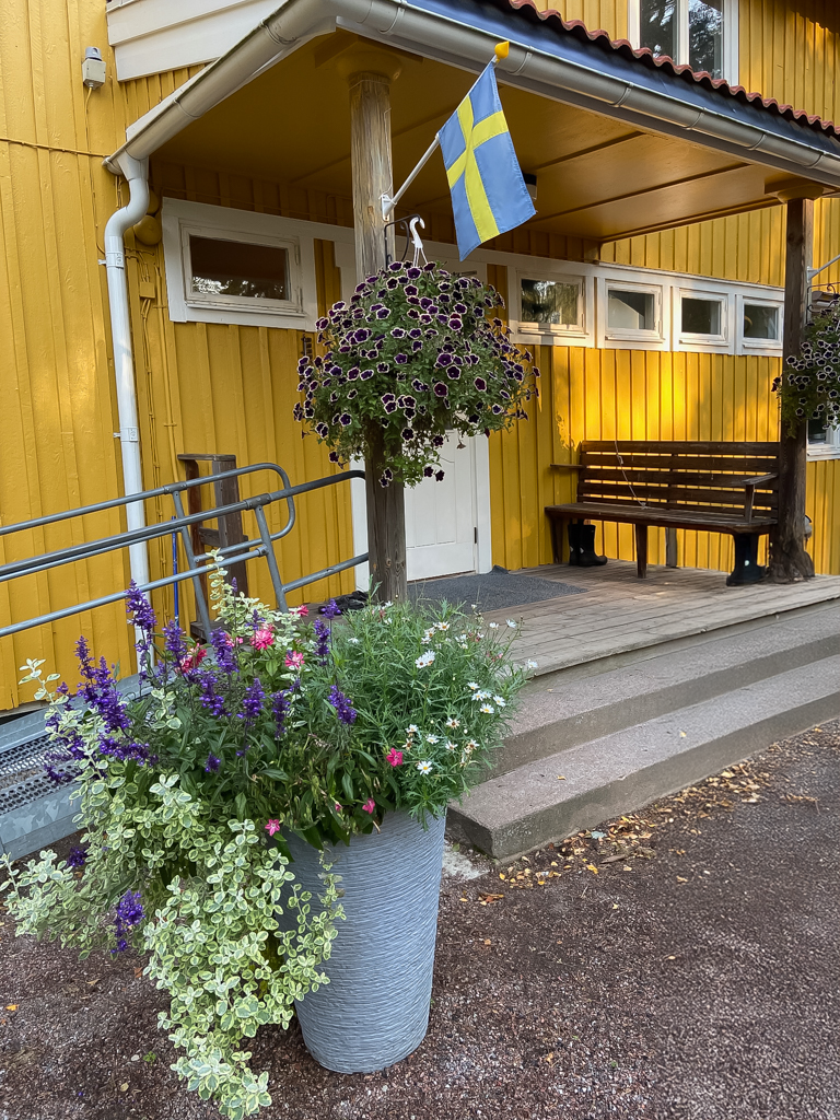 Gul byggnad med svenska flaggan och blommor vid entrén