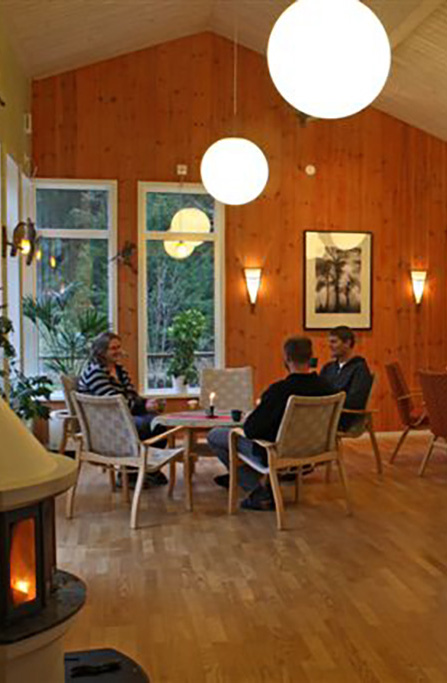 Möblerat samlingsrum med soffor, fåtöljer och belysning, där personer sitter och pratar.