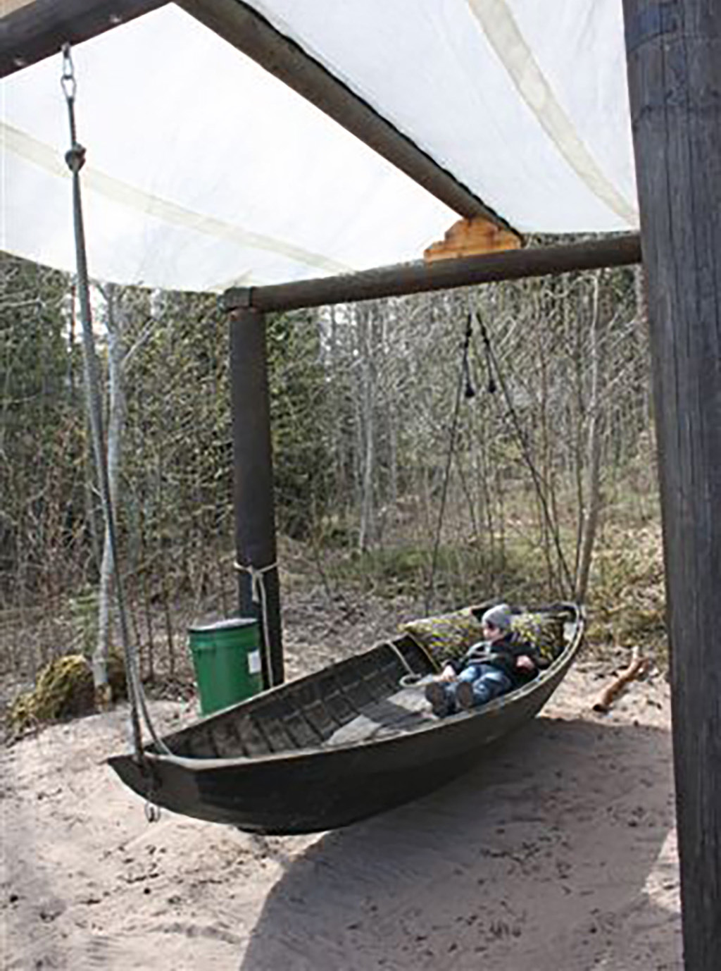Barn som gungar i en upphängd båt under ett tak i en skogsmiljö.